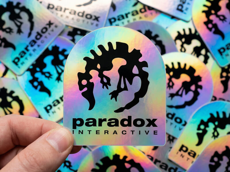 Hand som håller hologramklistermärke framför hög med tryckta dekaler i bulk