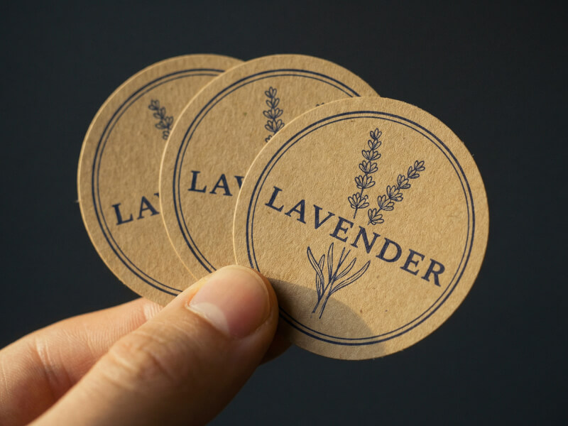 Hand som håller tre runda klistermärken i kraftpapper med lavendeldesign mot mörk bakgrund