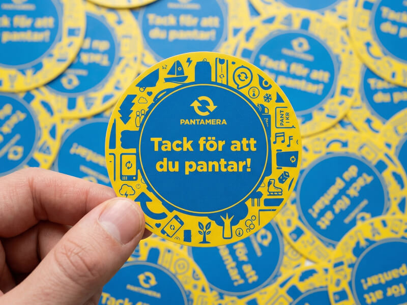 Hand som håller runt pappersklistermärke från Pantamera i gult och blått med återvinningsikoner och texten Tack för att du pantar
