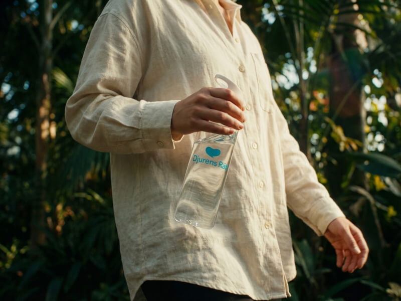 Person i beige linneskjorta som promenerar i en solbelyst inomhusträdgård hållandes en transparent Djurens Rätt vattenflaska