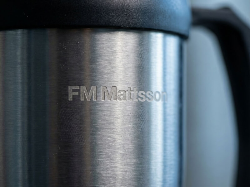 Närbild på en Sanibel termosmugg i borstat rostfritt stål med lasergraverad FM Mattsson-logotyp och svart lock.