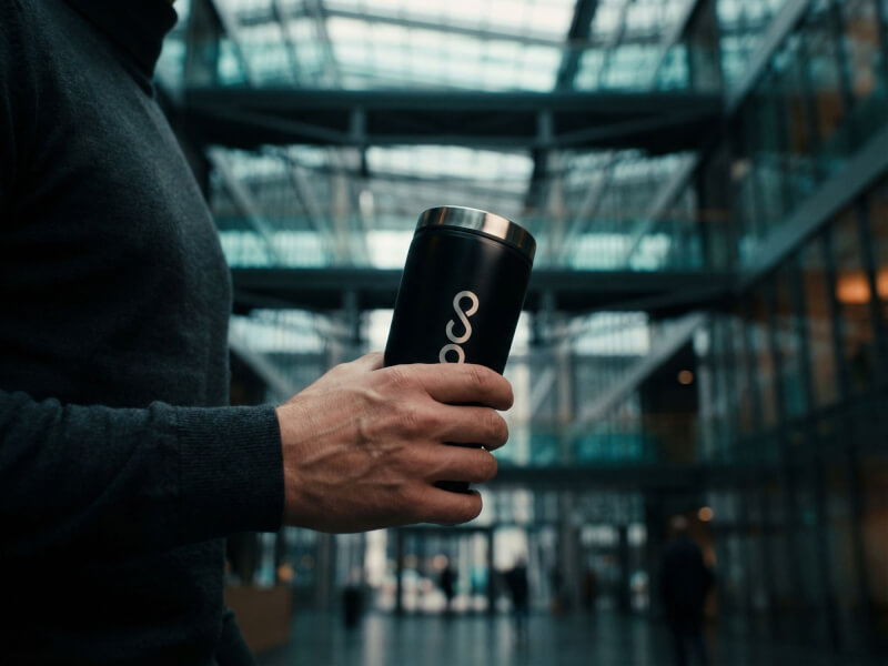 Person som håller i en svart matt termosmugg Elwood med laser-graverad logotyp i en modern kontorsmiljö med glasfasad.