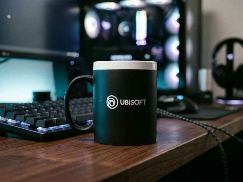 Svart matt kaffemugg Chalky med vit Ubisoft-logotyp placerad på ett skrivbord i en gamingmiljö med dator och tangentbord.