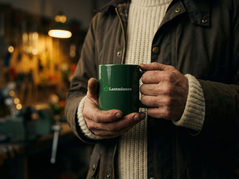 En person i en verkstadsmiljö håller en mörkgrön blank kaffemugg av modellen Santos med en tryckt Lantmännen-logotyp.