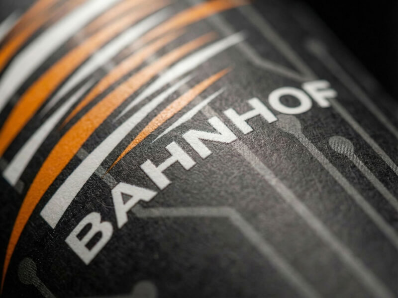 Närbild på en svart pappersmugg 35 cl med digitaltryckt logotyp för Bahnhof och tekniskt kretskortsmönster i orange och grått.