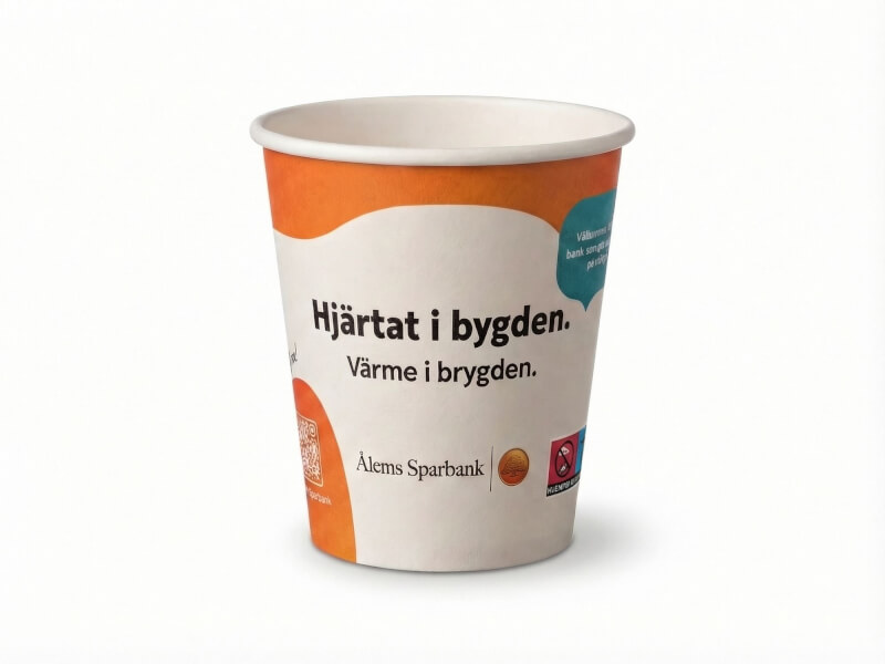 Enkelväggig pappersmugg på 24 cl med tryck för Ålems Sparbank mot vit bakgrund. Muggen har en matt finish med orangea detaljer och texten "Hjärtat i bygden".