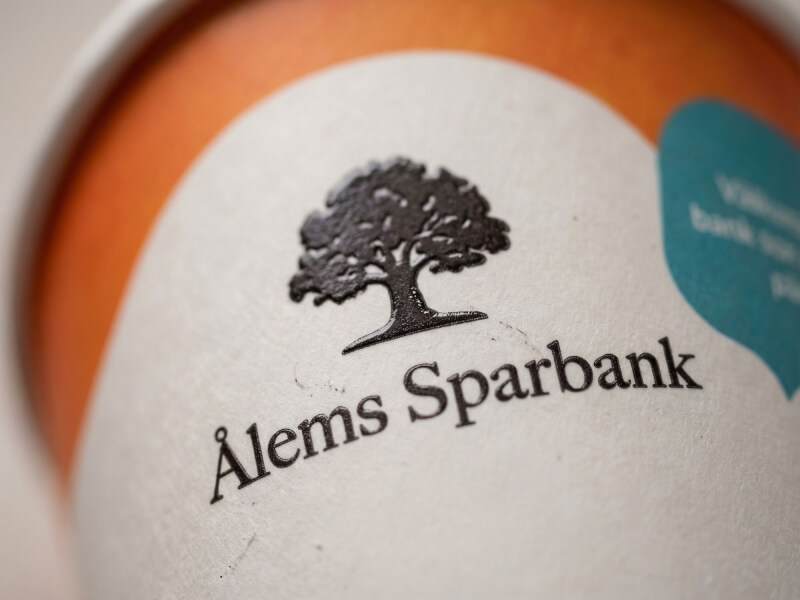 Närbild på en enkelväggig pappersmugg 24 cl med tryckt logotyp för Ålems Sparbank i svart färg på en matt, krämfärgad pappersyta.