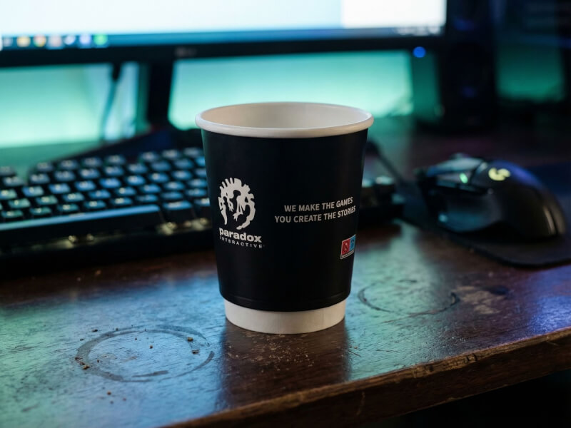 Dubbelväggig pappersmugg 24 cl med Paradox Interactive-logotyp placerad på ett skrivbord i en spelmiljö med tangentbord och datormus.