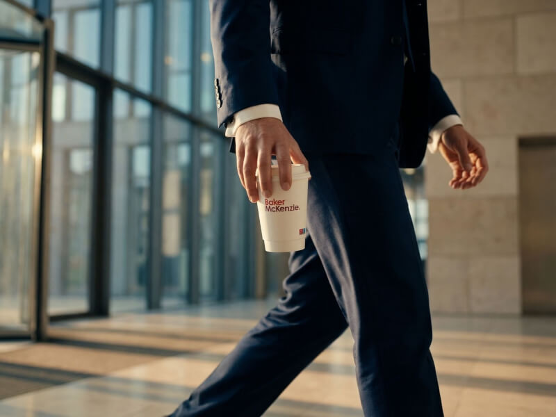 Affärsman i mörkblå kostym som går genom en modern kontorsbyggnad och håller i en vit pappersmugg med dubbla väggar (36 cl) med röd logotyp.