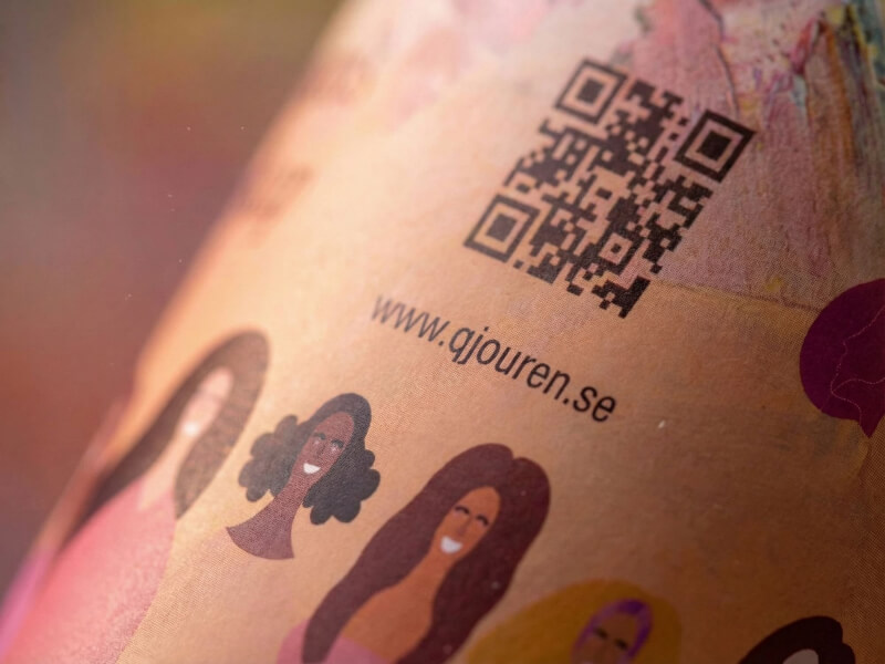 Närbild på en pappersmugg med dubbla väggar och 48 cl volym, med ett färgglatt digitaltryck föreställande illustrationer av människor, en QR-kod och webbadressen gjouren.se på en matt yta.