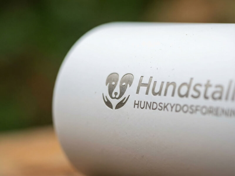 Närbild på ingraverad Hundstallet-logotyp på en matt vit metallflaska