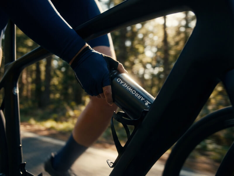 Cyklist med svart cykel på en vacker och solbelyst naturväg sträcker sig ner för att ta sin svarta Arrowhead-sportflaska ur sitt ställ