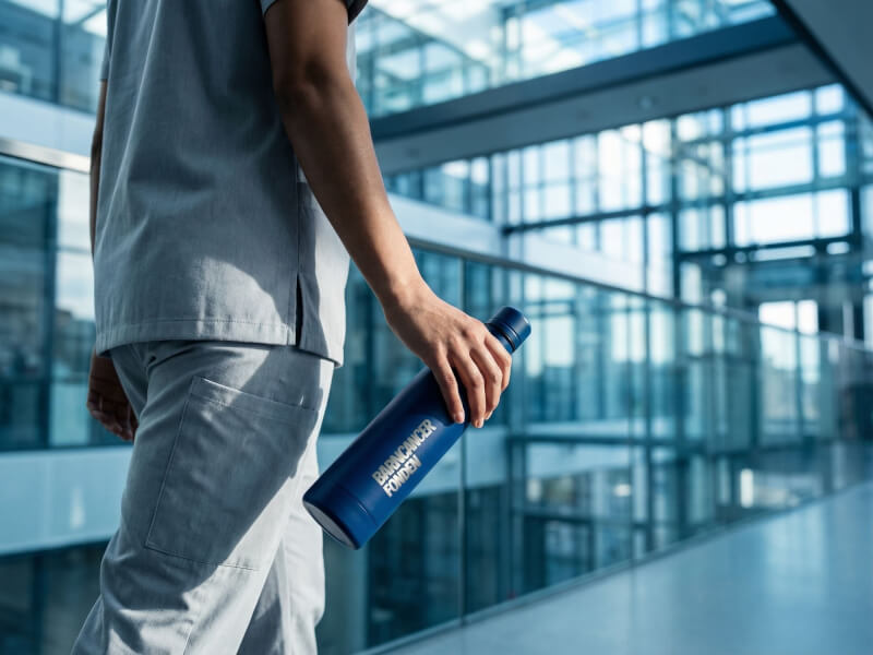En vårdpersonal i ljusgrå arbetskläder håller en matt mörkblå Termosflaska Vasa med silvrig lasergraverad logotyp i en ljus, modern sjukhusmiljö.