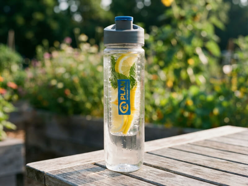 Transparent vattenflaska med Plan International-tryck fylld med skivad citron i infuserrör, stående på ett slitet träbord utomhus i trädgårdsmiljö