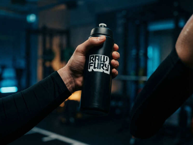 Två personer i mörka träningskläder där den ena överräcker en svart Raw Fury sportflaska i en mörkt belyst gymmiljö