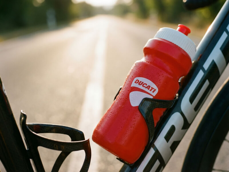 Röd Ducati sportflaska monterad i en cykels flaskställ intill en landsväg med varmt kvällsljus