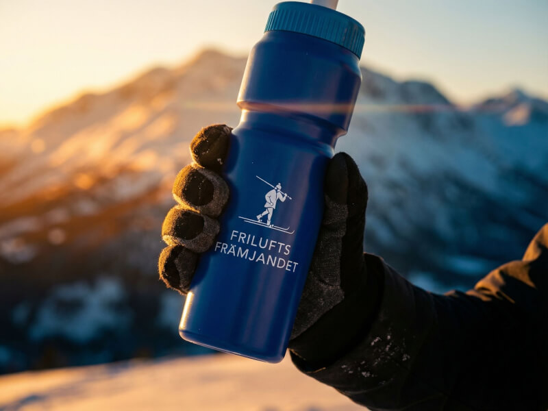 Behandskad hand lyfter en blå Friluftsfrämjandet-sportflaska i extrem motljus mot bakgrund av storslagna snötäckta bergstoppar vid solnedgång