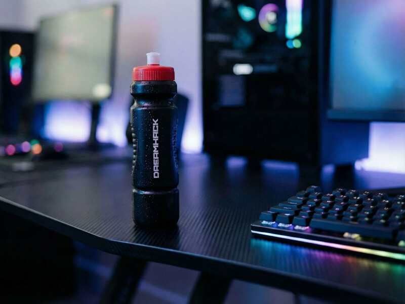 Svart Dreamhack sportflaska med rött lock stående på ett gamer-skrivbord upplyst av blått ljus från bildskärmar och LED