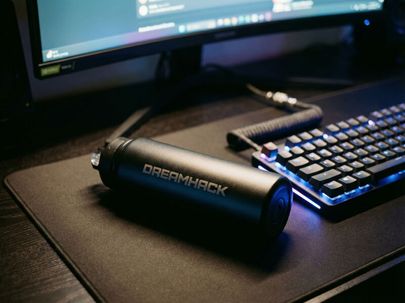 Svart sportvattenflaska med Dreamhack-tryck liggande på en svart skrivbordsmatta intill ett upplyst tangentbord i blått gaming-ljus