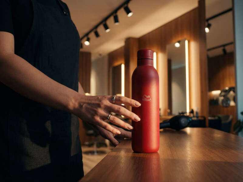 Matt röd Termosflaska Thor 510 med vitt tryck, stående på en träbänk i en frisersalong bredvid en frisör.