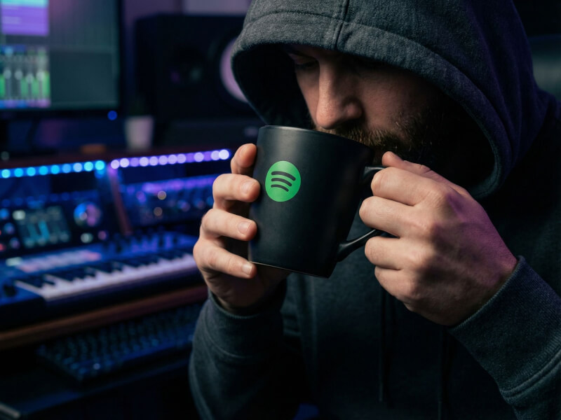 Person med huvtröja dricker kaffe ur en matt svart stengodsmugg Freja med grön Spotify-logotyp i en musikstudio.