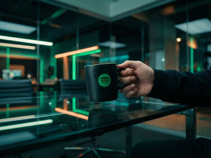 En person håller i en matt svart stengodsmugg med en tryckt grön Spotify-logotyp i ett modernt kontorslandskap med neonbelysning.