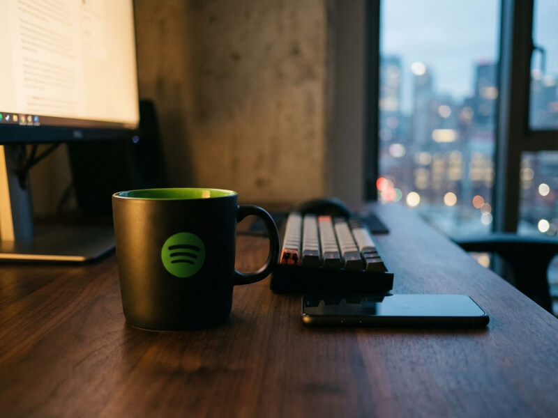 Svart stengodsmugg Emma med Spotify-logotyp placerad på ett skrivbord i en modern kontorsmiljö med utsikt över en stadssiluett.