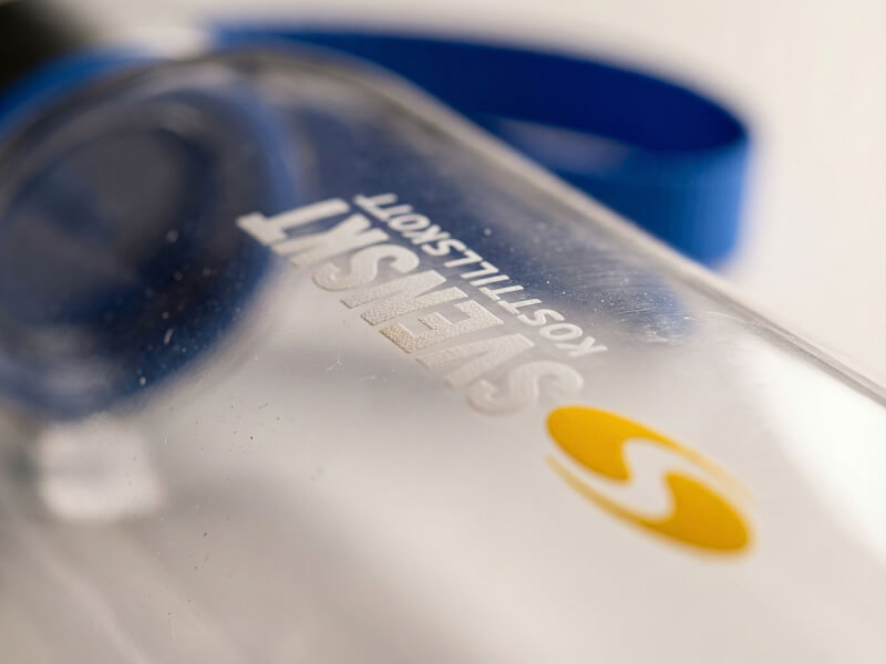 Närbild med kort skärpedjup av den vita och gula logotypen för Svenskt Kosttillskott tryckt på en transparent sportflaska