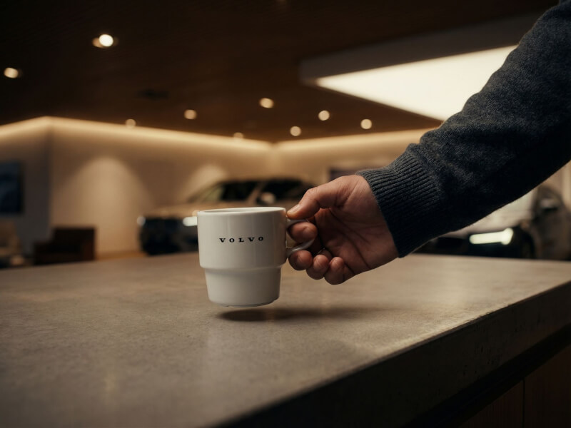 En person håller i en vit porslinsmugg Stina med Volvo-logotyp tryckt på sidan i en modern bilhall med dämpad belysning.