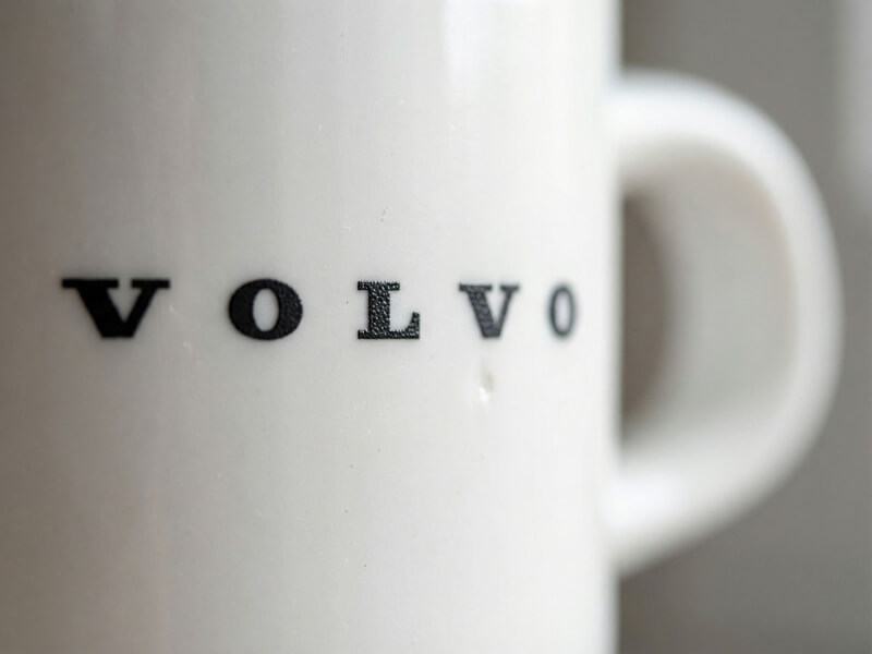 Närbild på en vit glansig porslinsmugg modell Stina med svart Volvo-logotyp tryckt på sidan.