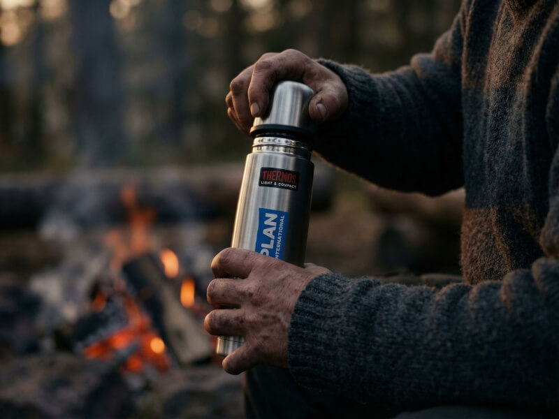 Person i ulltröja som utomhus en kylig kväll sitter vid en öppen, värmande lägereld och skruvar på locket på sin ståltermos