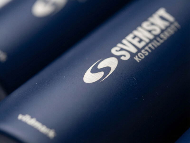 Skarpt vinklad närbild av vit och silverfärgad logotyp för Svenskt Kosttillskott tryckt på en matt mörkblå termos