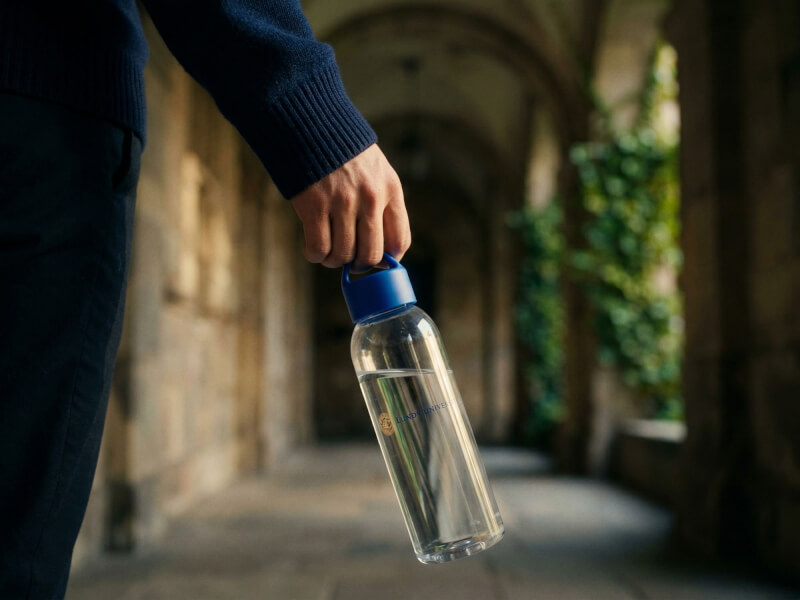 Hand i mörkblå tröja bär en transparent vattenflaska med mörkblått lock och Lunds Universitet-tryck längs ett stengalleri eller valvgång