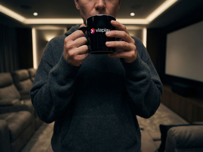Person som dricker ur en blank svart stengodsmugg med tryckt Viaplay-logotyp i ett hembiovarum