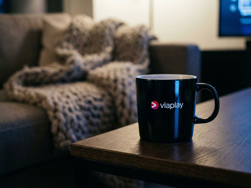 Svart blank stengodsmugg Emma med tryckt Viaplay-logotyp placerad på ett soffbord i en hemtrevlig vardagsrumsmiljö.