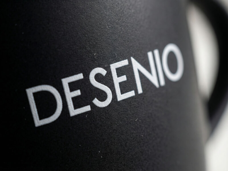 Närbild på en matt svart kaffemugg Riviera med vit tryckt logotyp för Desenio, som visar den högkvalitativa trycktekniken.