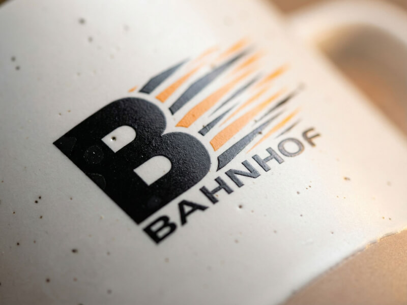 Närbild på en beige keramisk kaffemugg av modellen Pascal med en högkvalitativ screentryckt Bahnhof-logotyp i svart och orange.