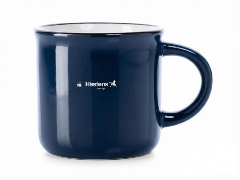 Mörkblå blank kaffemugg i vintagestil med vitt Hästens-tryck, isolerad mot vit bakgrund.