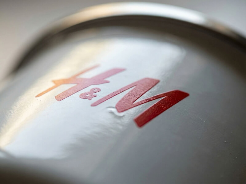 Närbild av en vit emaljmugg av modellen Kaffemugg Camp med en röd H&M-logotyp tryckt på den glansiga ytan.