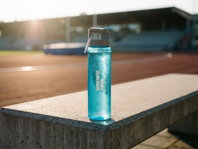 Ljusblå transparent sportflaska från Barncancerfonden placerad på en låg stenmur vid friidrotts- eller löparbanan i kvällsljus
