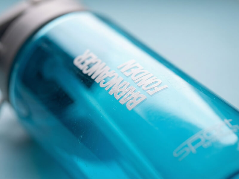 Närbildsdynamisk vinkel av vitt glänsande tryck med texten Barncancerfonden på den transparenta ljusblå sportflaskan