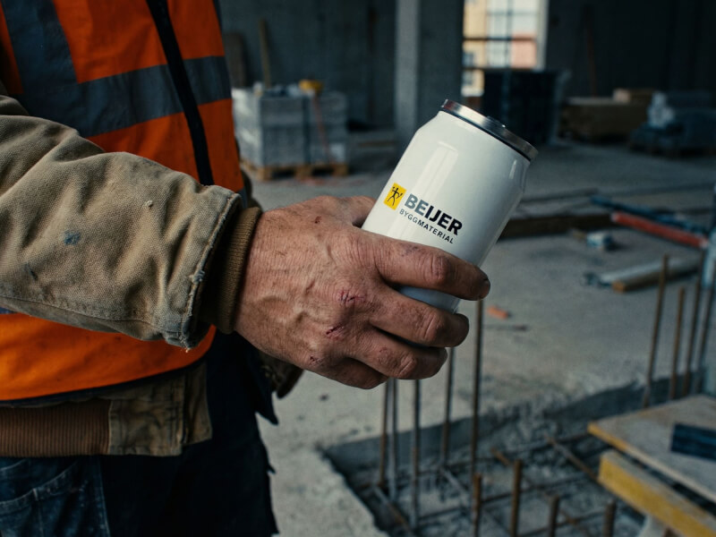En arbetare på en byggarbetsplats håller i en vit termosmugg Can 280 med Beijer Byggmaterial-logotyp tryckt på sidan.