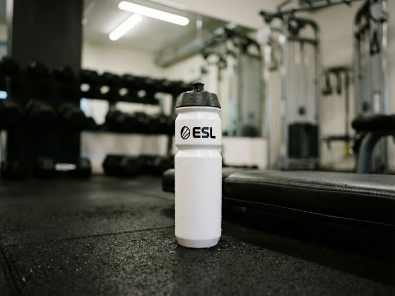 Vit ESL-sportflaska stående på golvmattan i ett modernt gym med hantlar och träningsbänk i bakgrunden