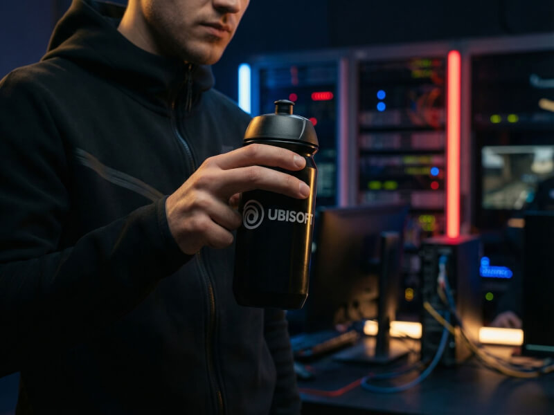 Person i svart tröja som i ett rum intill upplysta servrar håller i en svart sportflaska med vit Ubisoft-logotyp