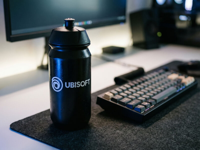 Svart Ubisoft-sportflaska stående på en svart skrivbordsunderlägg intill ett datortangentbord i en dämpat belyst datormiljö