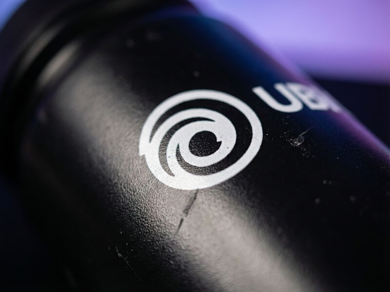 Skarp närbild på Ubisofts ikoniska vita swirl-logotyp tryckt på en mörkt texturerad sportflaska