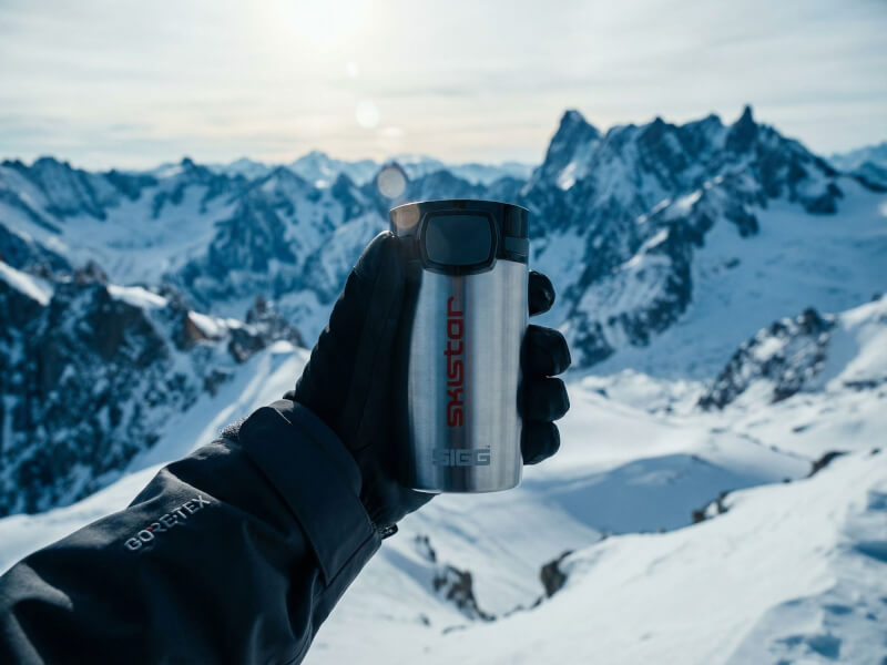 En person i skidhandskar håller en silverfärgad rostfri termosmugg Miracle med Skistar-logotyp i rött tryck framför ett snöigt alpint berglandskap.