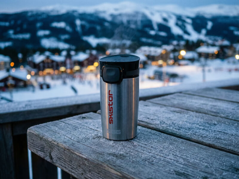 Termosmugg Miracle i rostfritt stål med röd SkiStar-logotyp tryckt på sidan, placerad på ett frostigt träbord med en snöig skidort i bakgrunden.