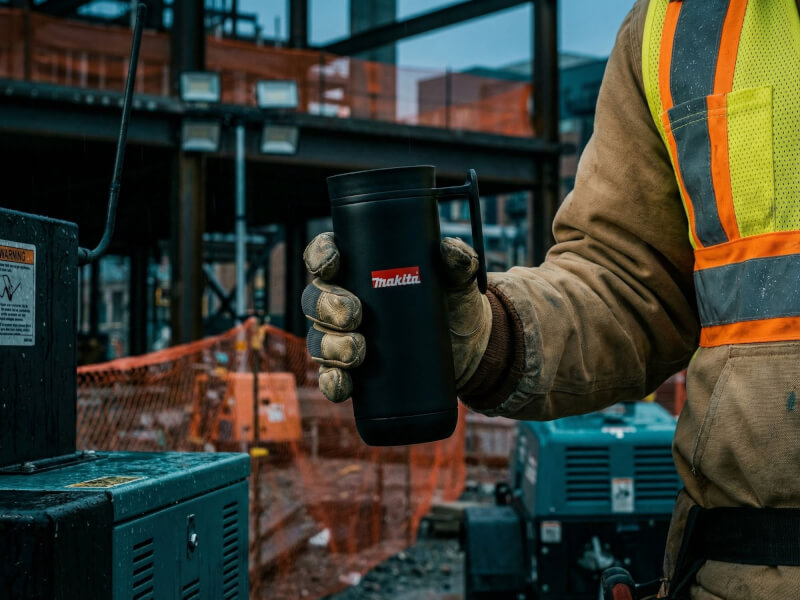Svart matt termosmugg Clik med Makita-logotyp hållen av en byggarbetare i skyddskläder på en byggarbetsplats.