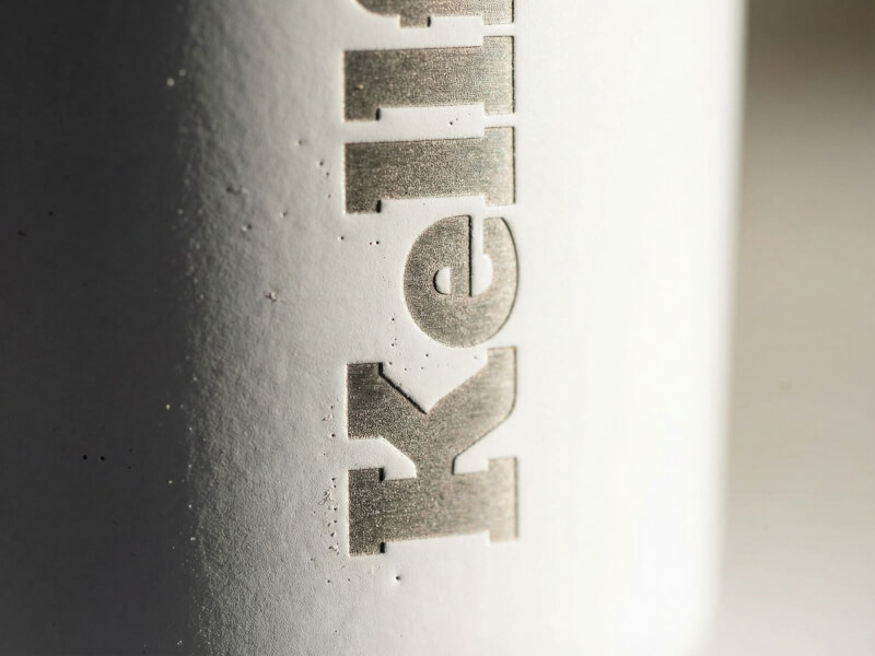 Närbild av en vit termosmugg Mini med logotyp utförd i precisionslasergravyr som blottar den metalliska ytan under den matta beläggningen.